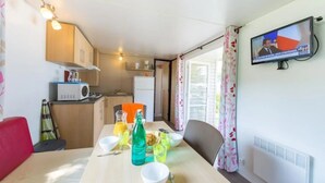 A compact room with a dining table set for two, a kitchen area, a television mounted on the wall, and a window with curtains. - ZEcamping 3 étoiles animaux acceptés avec piscine chauffée à Guérande (Guérande)