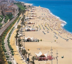 Una playa con filas de tiendas de campaña y sombrillas, un paseo marítimo con palmeras y edificios al fondo.
