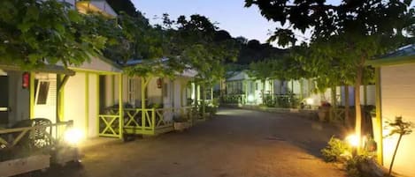 A row of single-story cabins with a dirt driveway, surrounded by trees and lit by warm lights.