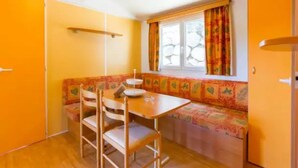 A compact kitchen with a wooden table and chairs, a bench with a floral cushion, and a window with curtains.