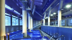 A modern indoor swimming pool with clear water, a central water feature, and a staircase leading to upper levels.