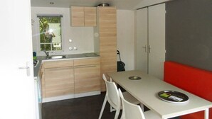 A compact kitchen with a dining area, a red bench, and a window with blinds. - ZEcamping 3 étoiles piscine mobil-home TV 29m2 5 pers (Urt)