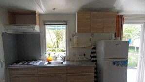 Eine kleine Küche mit Gasherd, Spüle und einem weißen Kühlschrank. Ein Fenster bietet Blick auf Grünflächen.