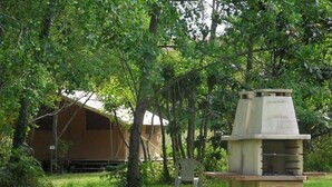 A campsite with a concrete barbecue grill, a tent, and a chair in a grassy area surrounded by trees. - ZEcamping 3 étoiles piscine pet friendly bien-être locatif TV 22m2 4 pers (Urt)