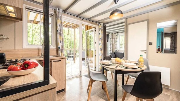 A modern kitchen with a dining area, featuring a large window, a dining table with chairs, and a bowl of fruit on the counter. - ZEcamping 4 étoiles espace aquatique chalet TV BBQ clim 35m2 (Loupiac)