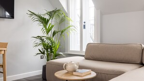 A modern living room with a beige sofa, a round wooden table with a teapot and book, and a large green plant. - One Bedroom Apartment, Sleeps 2 (London)