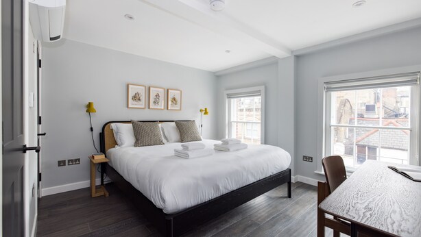 A modern bedroom with a large bed, bedside tables, a dining table, and a window with a city view. - One Bedroom Apartment, Sleeps 2 (London)