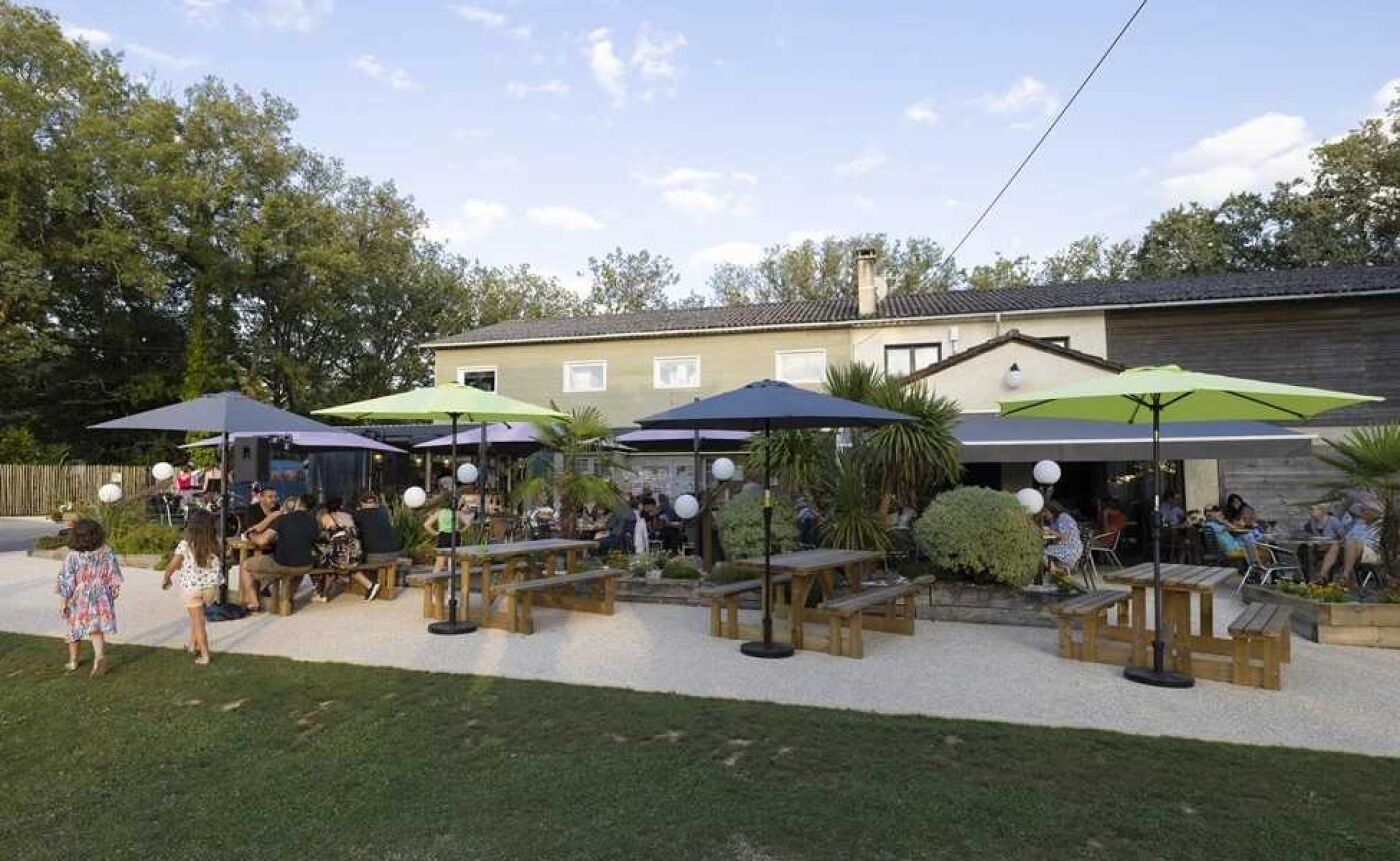 Ein Außenbereich mit einer Terrasse, Holztischen und grünen Sonnenschirmen, umgeben von einem Gebäude mit einem Ziegeldach.