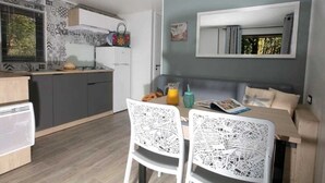 A modern kitchen with grey cabinets, a white countertop, and a built-in oven. There is a dining area with a table and chairs, and a window with blinds. - ZEcamping 4 étoiles parc aquatique club enfant mobil-home TV BBQ 26m2 4 pers (Loupiac)