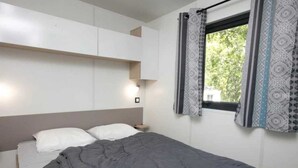 A bedroom with a bed, a bedside table, a wall-mounted shelf, and a window with patterned curtains. - ZEcamping 4 étoiles parc aquatique club enfant mobil-home TV BBQ 26m2 4 pers (Loupiac)