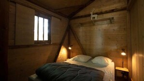 Um quarto de sótão acolhedor, com teto inclinado de madeira, uma cama simples com roupa de cama branca e duas luminárias fixadas na parede.