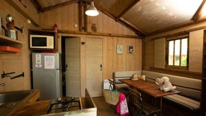 Interior rústico de uma cabana, com paredes de madeira, cozinha compacta com micro-ondas e frigorífico, um banco e uma janela.