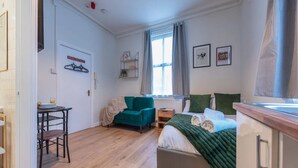 Quarto de hotel compacto com cama, sofá, mesa pequena e cadeira. Há uma janela com cortinas, um radiador e uma prateleira fixada na parede com objetos.