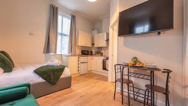 Um espaço compacto com cama, sofá verde, cozinha compacta e TV fixada na parede.
