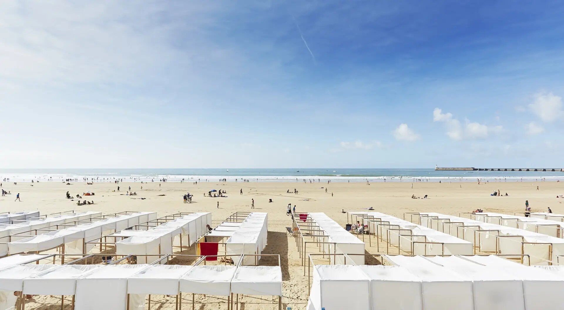 Une plage bordée de rangées de tentes blanches, avec une jetée visible au loin.