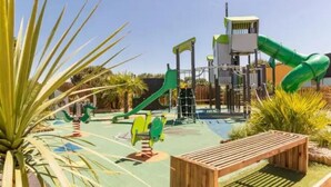 A playground with various play structures, including slides and climbing areas, surrounded by palm trees and wooden benches.