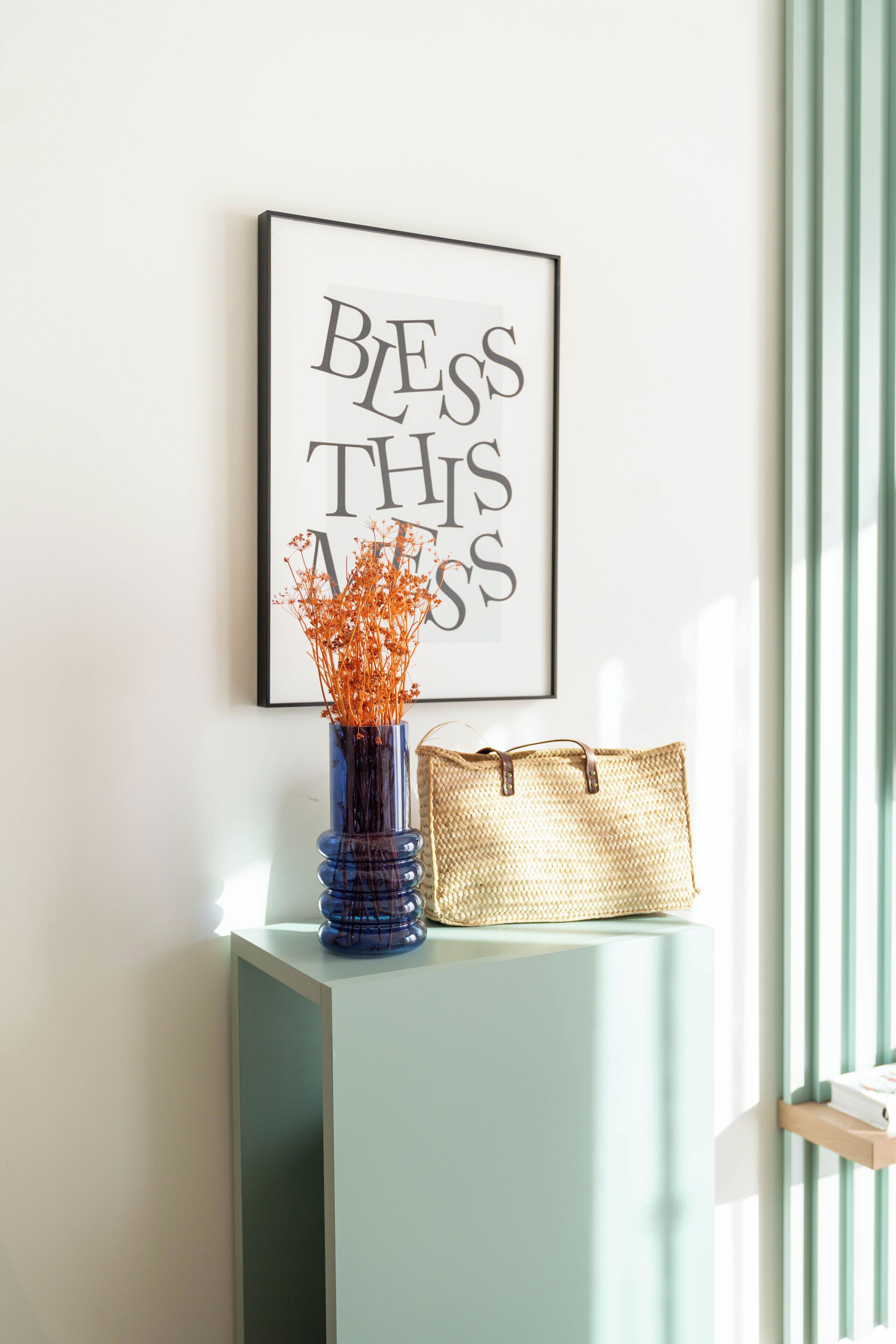 A framed inspirational quote on a wall, a blue vase with dried flowers, and a woven bag on a light teal console table.