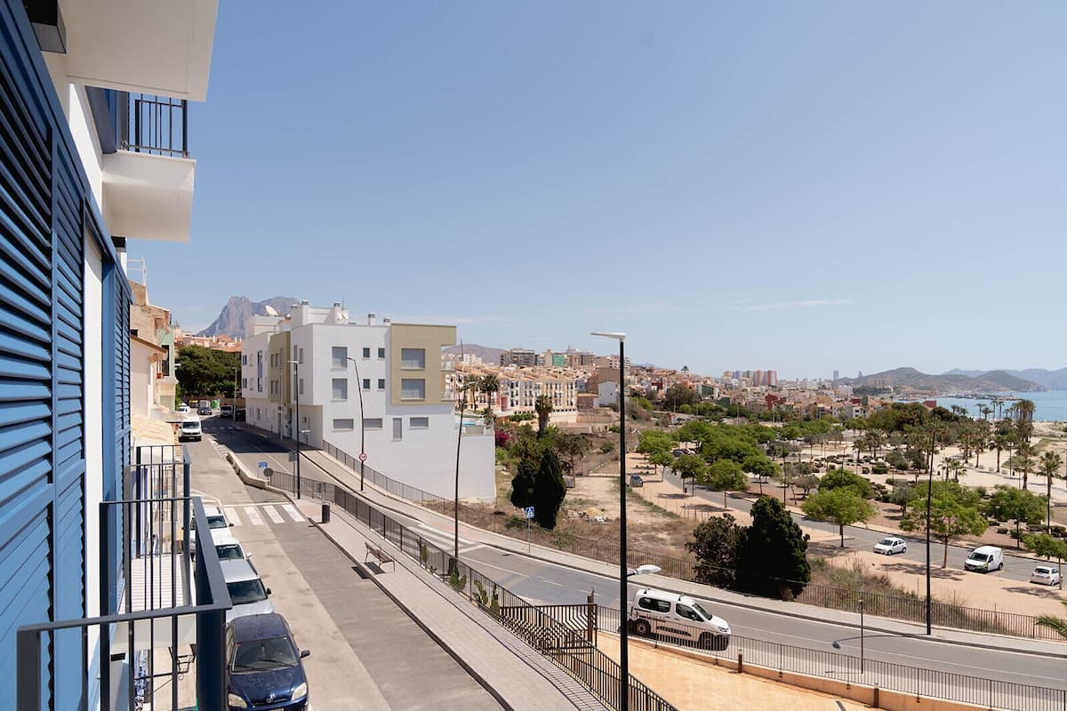 A coastal cityscape with modern buildings, a clear sky, and a road with vehicles.
