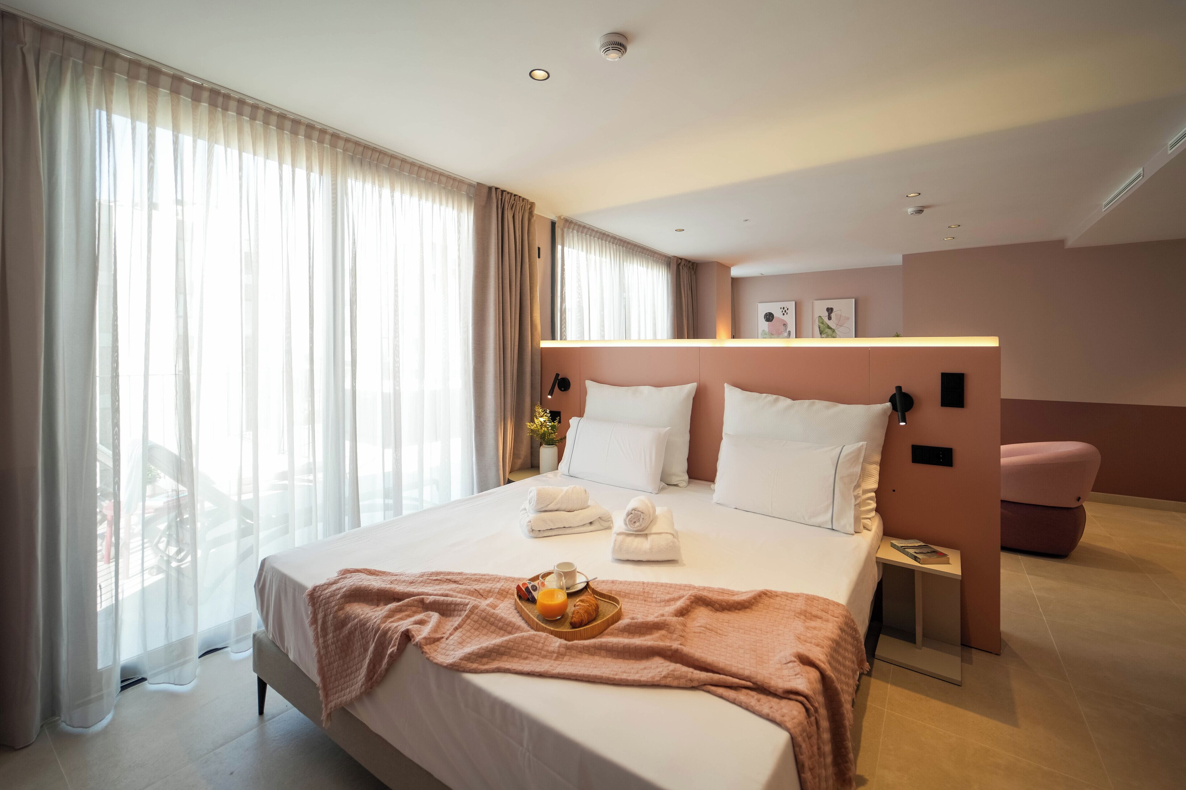 Ett modernt hotellrum med en stor säng, ett nattduksbord med en lampa, en rosa stol och ett frukostbricka på sängen.