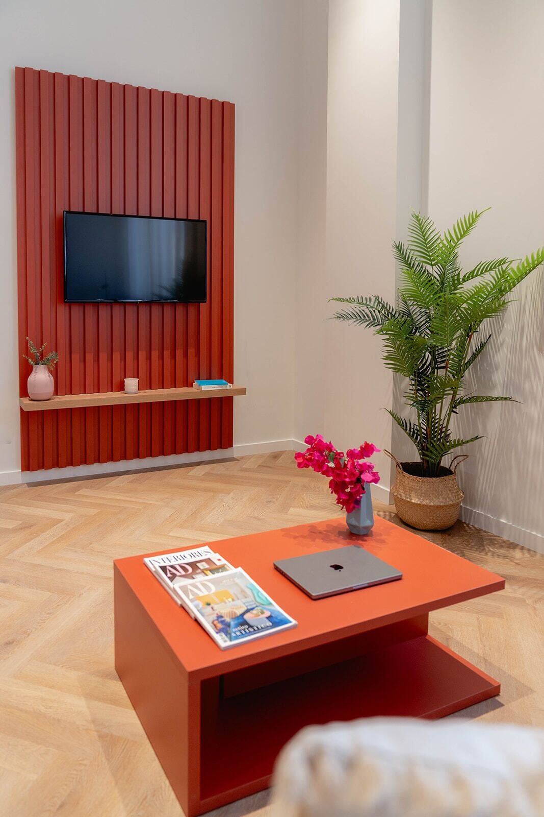 Una sala de estar moderna con paneles de pared rojos, un televisor de pantalla plana montado sobre una repisa de madera, una mesa de centro con una computadora portátil y revistas, y una planta verde grande.