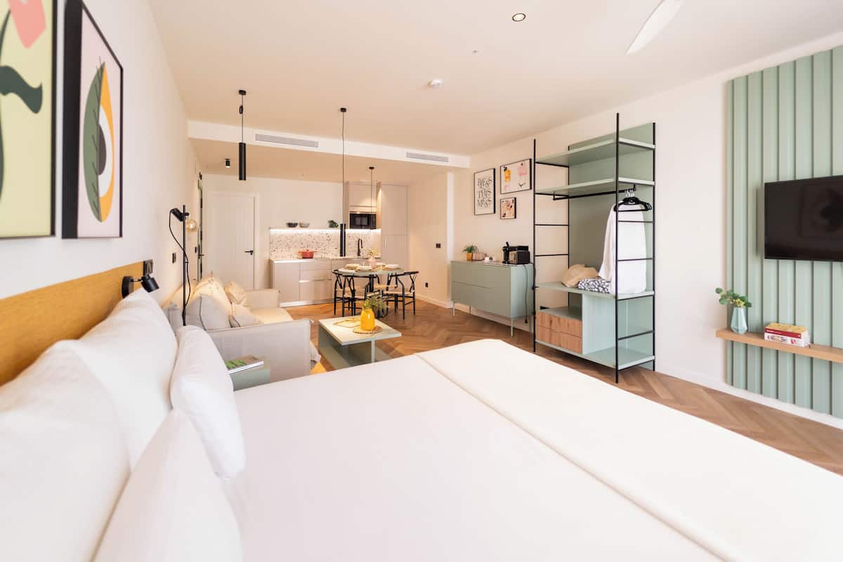 A modern hotel room with a large bed, a dining area, kitchenette, and a TV mounted on the wall.