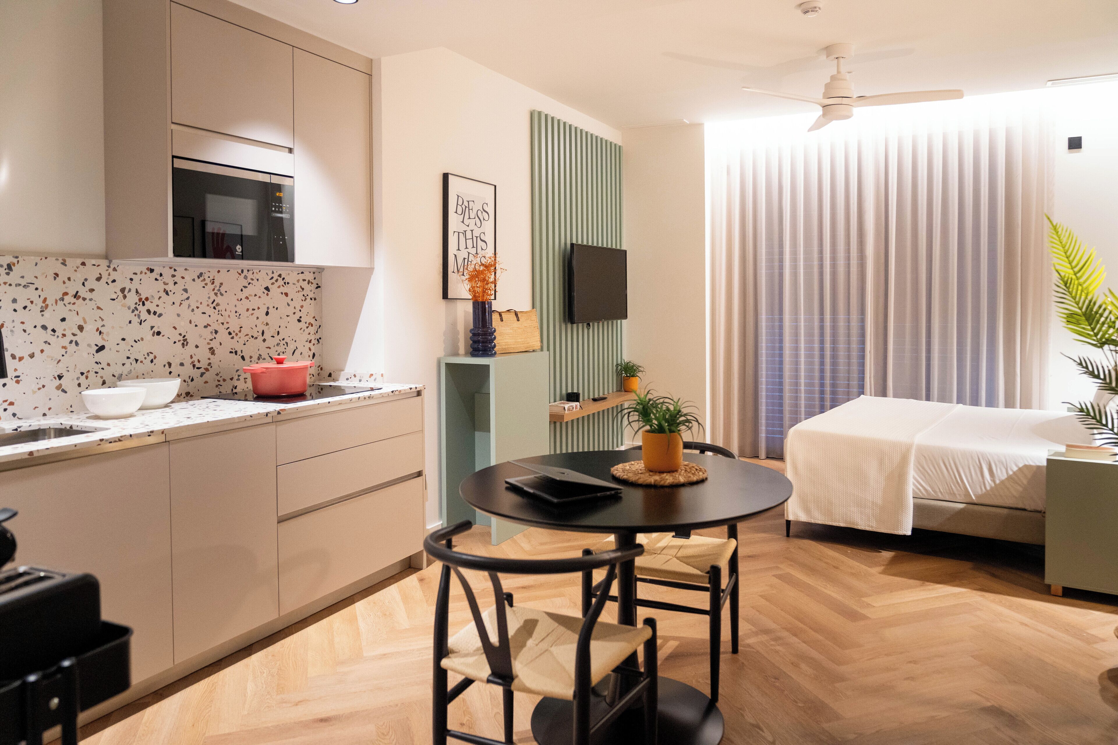 A modern kitchen with a microwave, sink, and countertop. There is a dining table with chairs and a small potted plant. A bed with a white blanket is visible in the background.