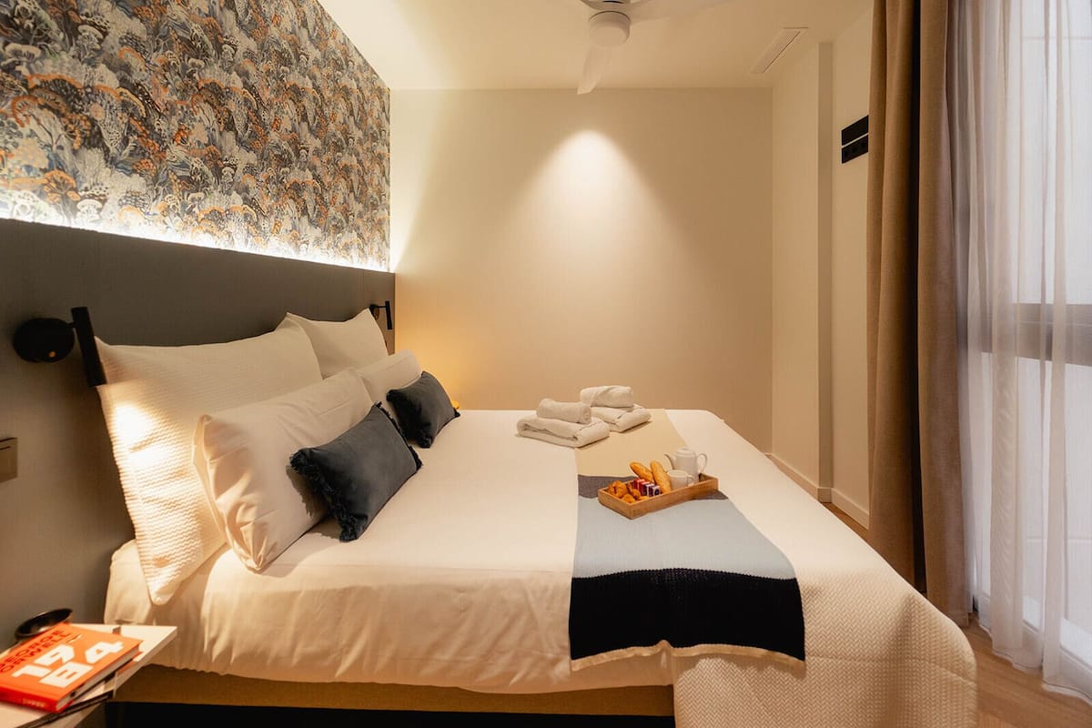 A neatly made bed with pillows, a tray of breakfast, and a book on the bedside table.
