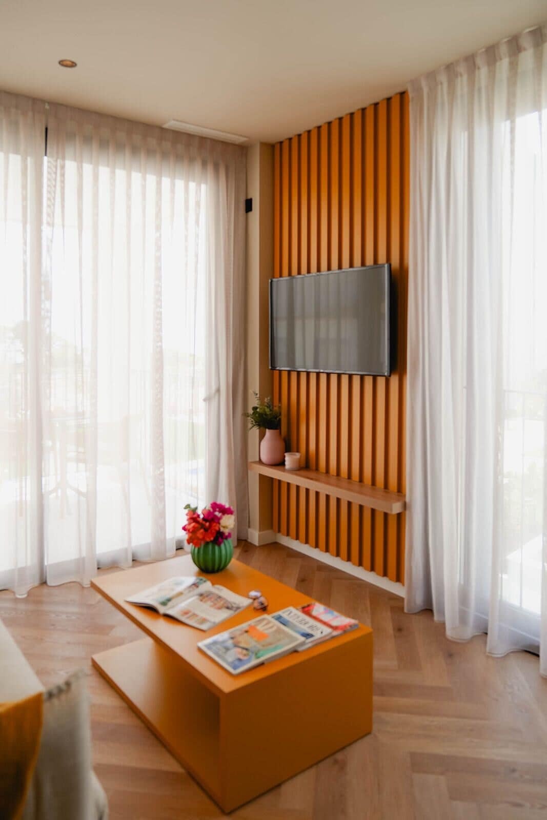 Een moderne woonkamer met een flatscreen televisie bevestigd aan een houten wandpaneel, een bijzettafeltje met tijdschriften, en een vaas met bloemen.