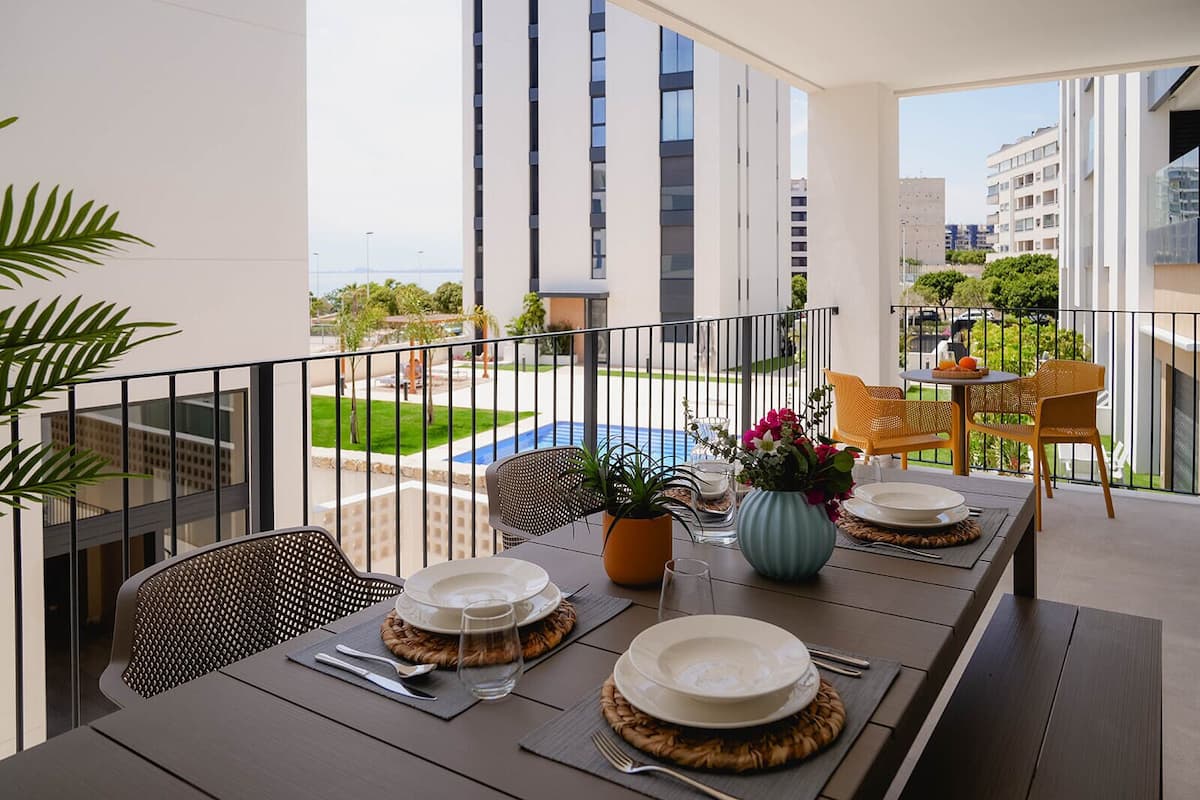 A balcony with a dining table set for two, featuring a view of a swimming pool and surrounding buildings.