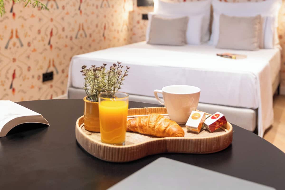 A neatly made bed with white linens, a bedside table with a tray of breakfast including croissant, orange juice, and tea, and a vase with flowers.