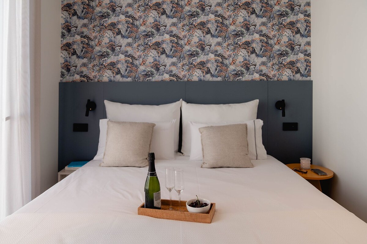 A hotel room with a large bed, two pillows, a nightstand with a phone and a glass, and a wall with a floral pattern.