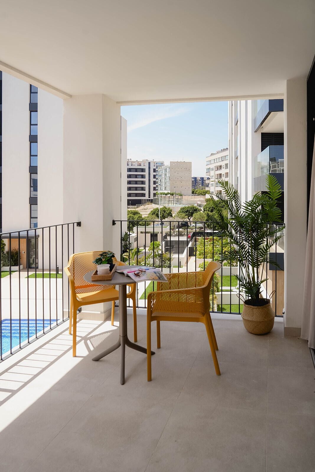 A balcony with a table and two chairs, a potted plant, and a view of a swimming pool and buildings.