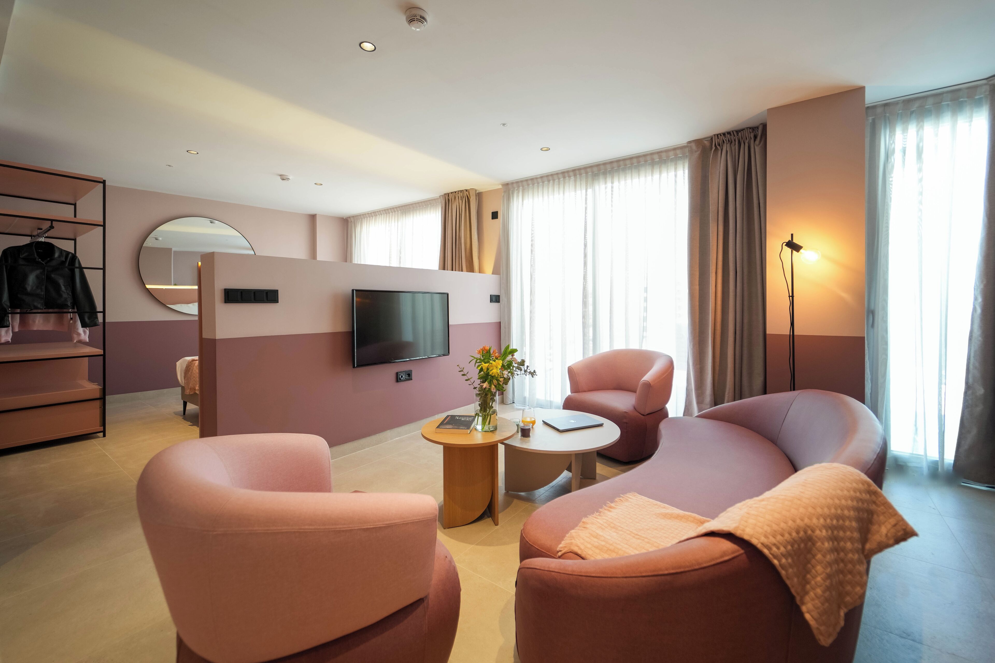A modern hotel room with a sofa, armchairs, a TV, a small table with a vase, and a wardrobe.