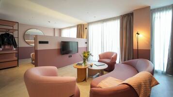 A modern hotel room with a sofa, armchairs, a TV, a small table with a vase, and a wardrobe.