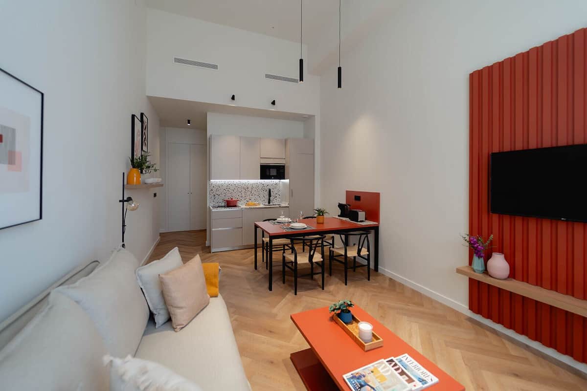 A modern apartment with a living room, dining area, and kitchen. There is a sofa, a dining table with chairs, a coffee table, and a TV mounted on the wall.