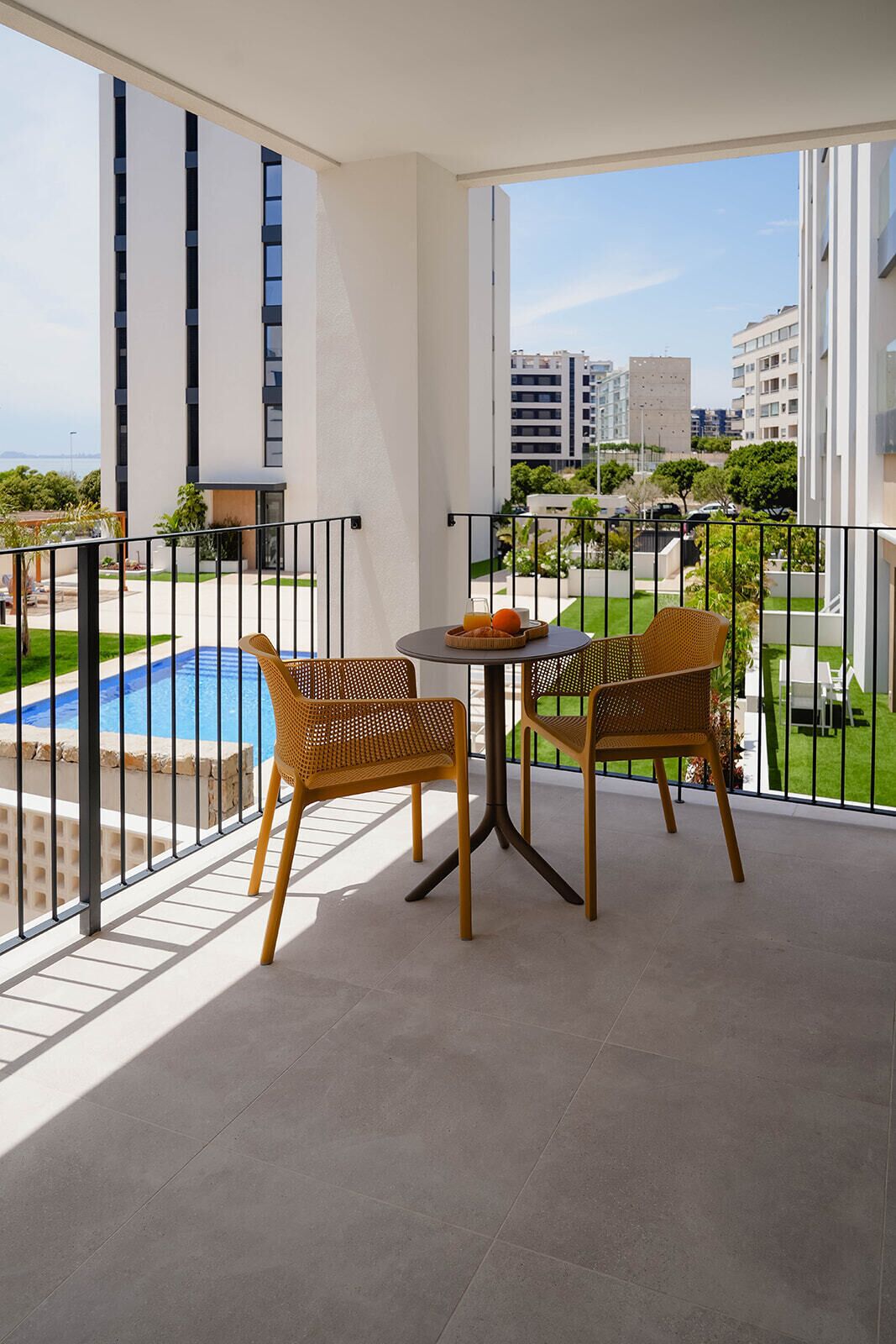 A balcony with wicker chairs and a table, overlooking a swimming pool and greenery.