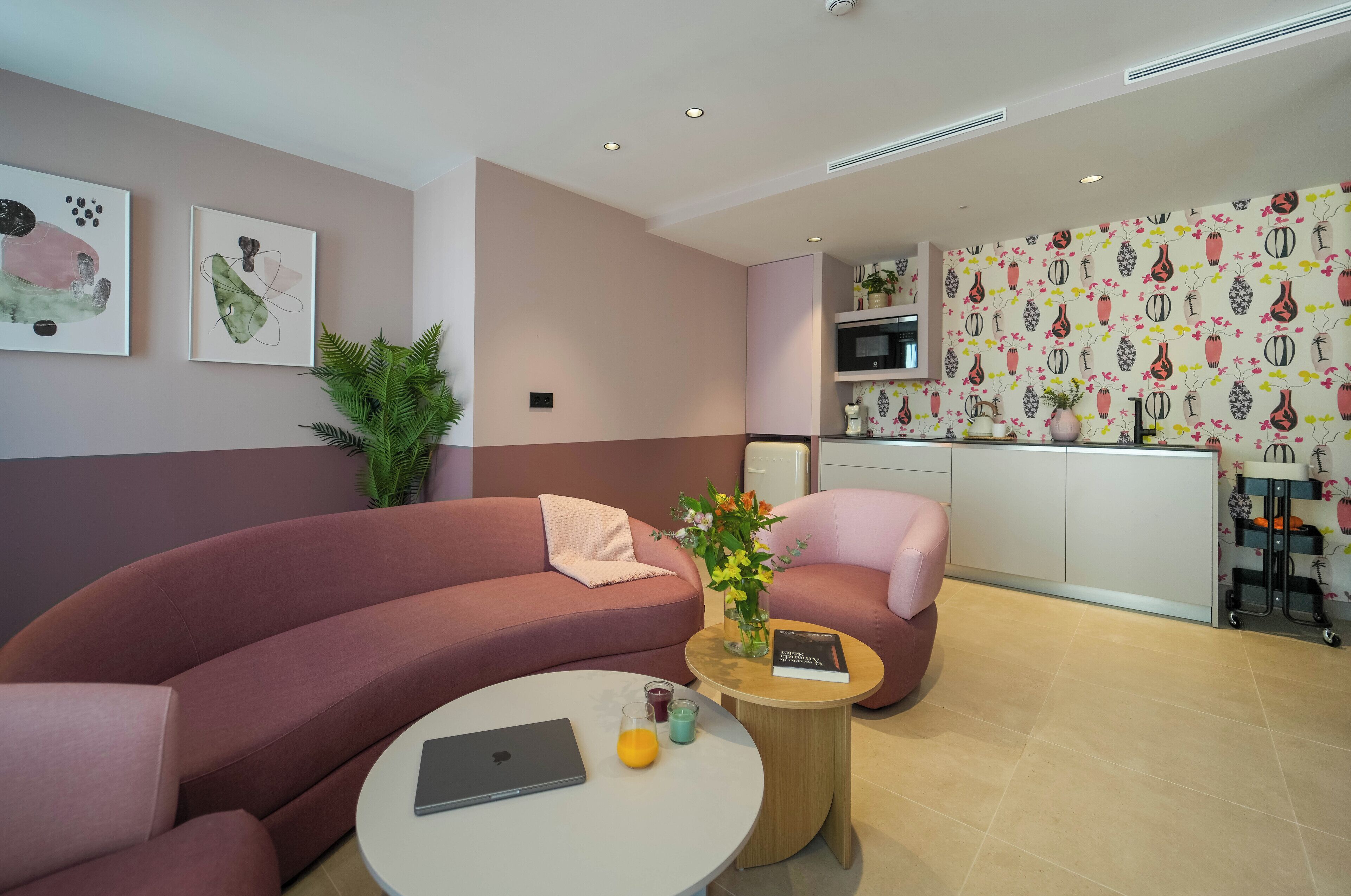 Una sala de estar moderna con un sofá curvo, una mesa de centro redonda con una computadora portátil y bebidas, y un sillón rosa.