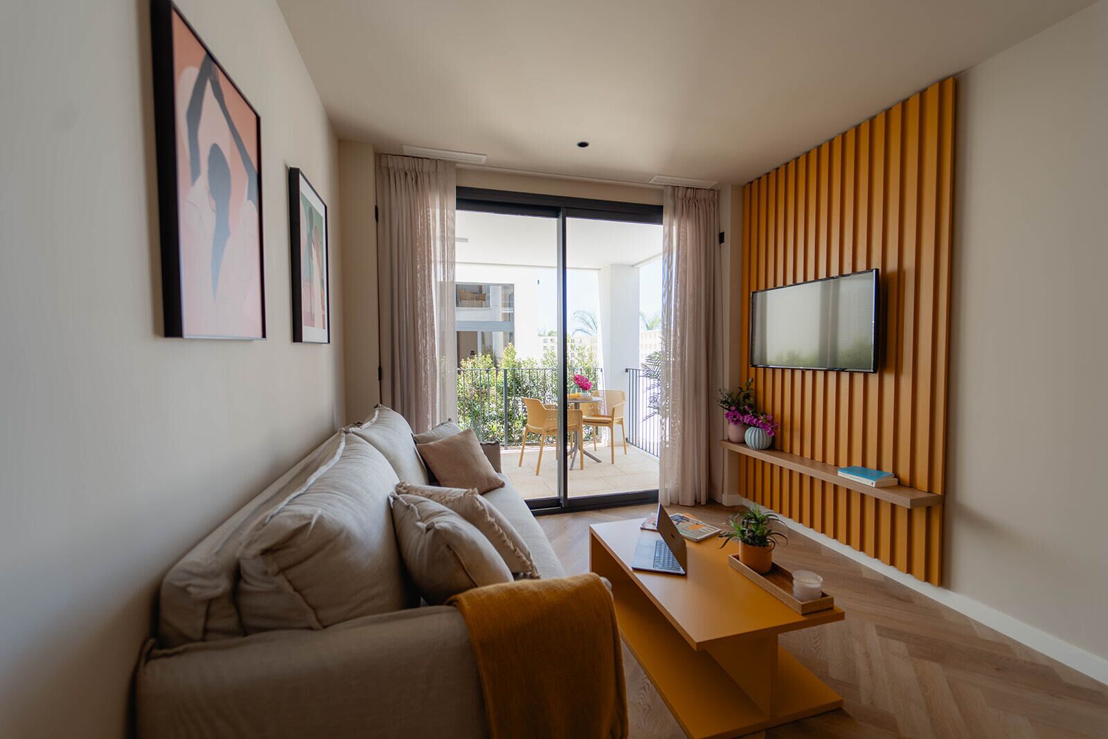 Una sala de estar moderna con un sofá, una mesa de madera con una computadora portátil, un televisor montado en un panel de madera y un balcón con mobiliario exterior.