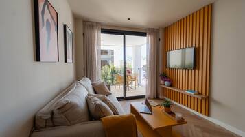 Una sala de estar moderna con un sofá, una mesa de madera con una computadora portátil, un televisor montado en un panel de madera y un balcón con mobiliario exterior.