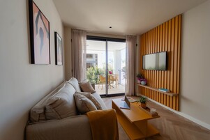 Una sala de estar moderna con un sofá, una mesa de madera con una computadora port átil, un televisor montado en un panel de madera y un balcón con mobiliario exterior.