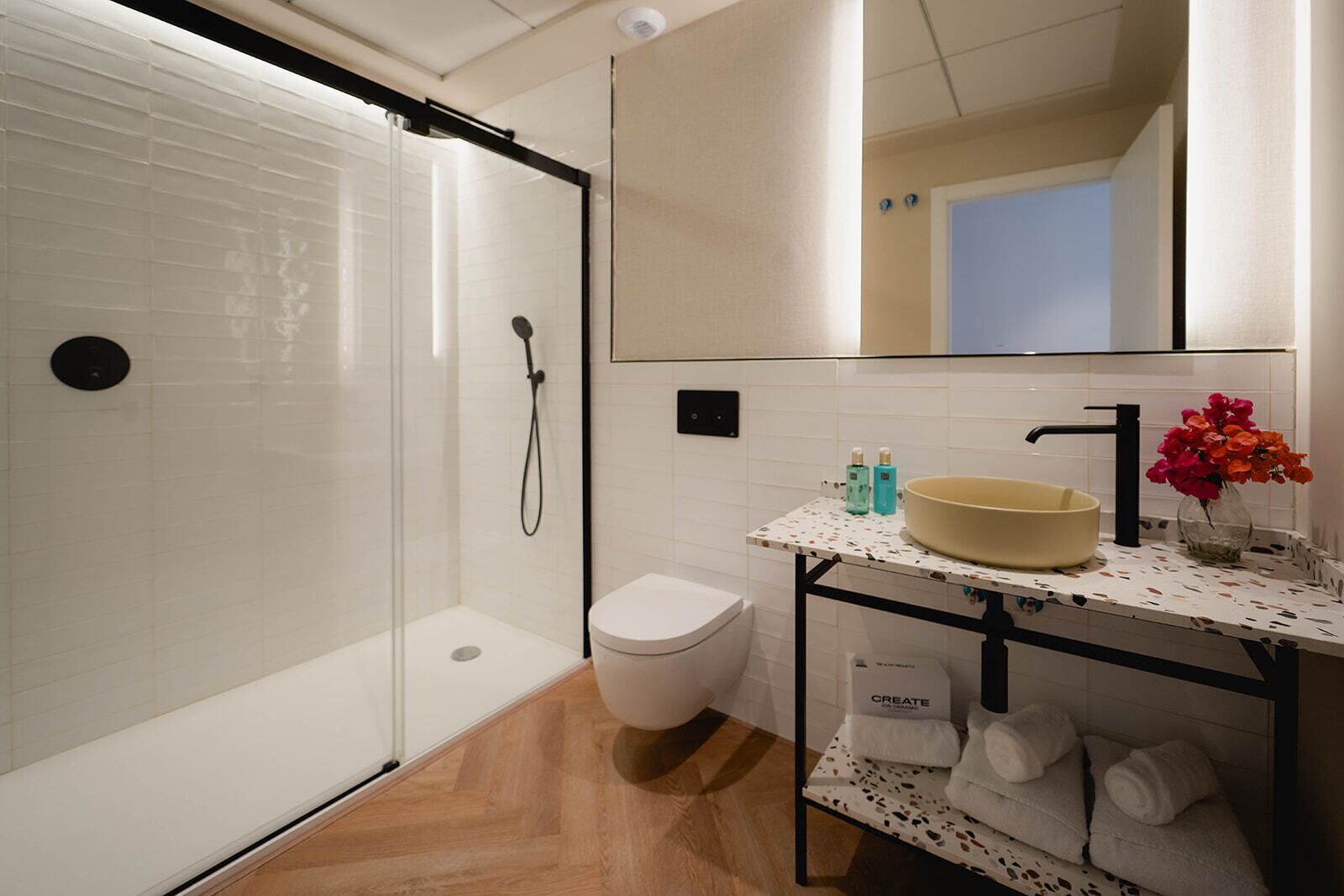 Une salle de bain moderne avec une douche à parois de verre, des toilettes blanches, un lavabo sur pied avec un receveur beige et un miroir au-dessus du lavabo.
