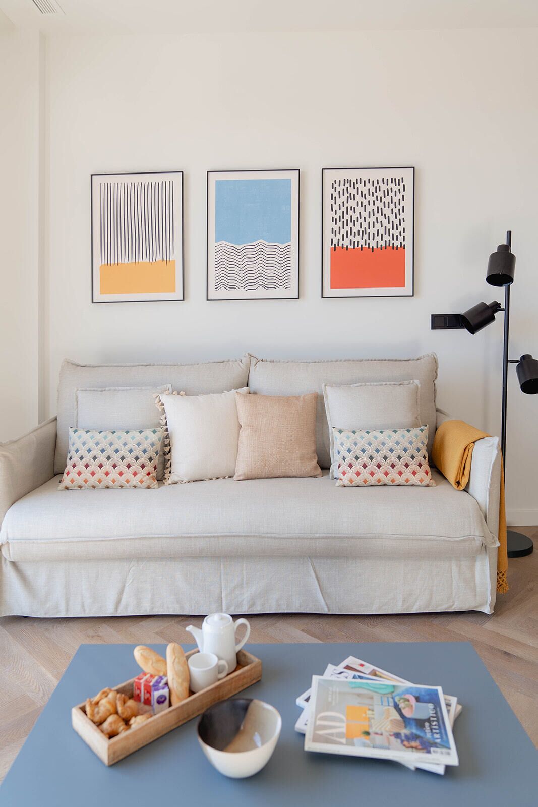 A living room with a sofa, pillows, a coffee table with snacks and magazines, and three framed artworks on the wall.