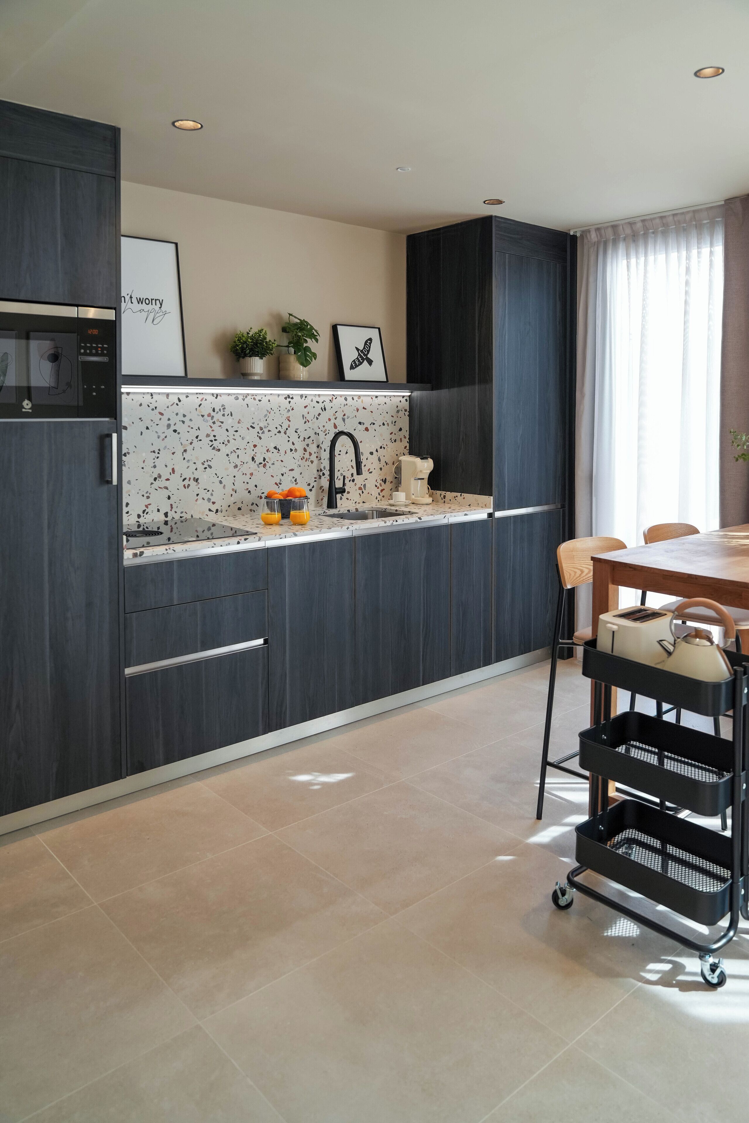 A modern kitchen with dark cabinetry, a white speckled backsplash, and a countertop with a sink and a kettle. There is a dining area with a wooden table and chairs, and a black cart with baskets.