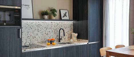 A modern kitchen with dark cabinetry, a white speckled backsplash, and a countertop with a sink and a kettle. There is a dining area with a wooden table and chairs, and a black cart with baskets.