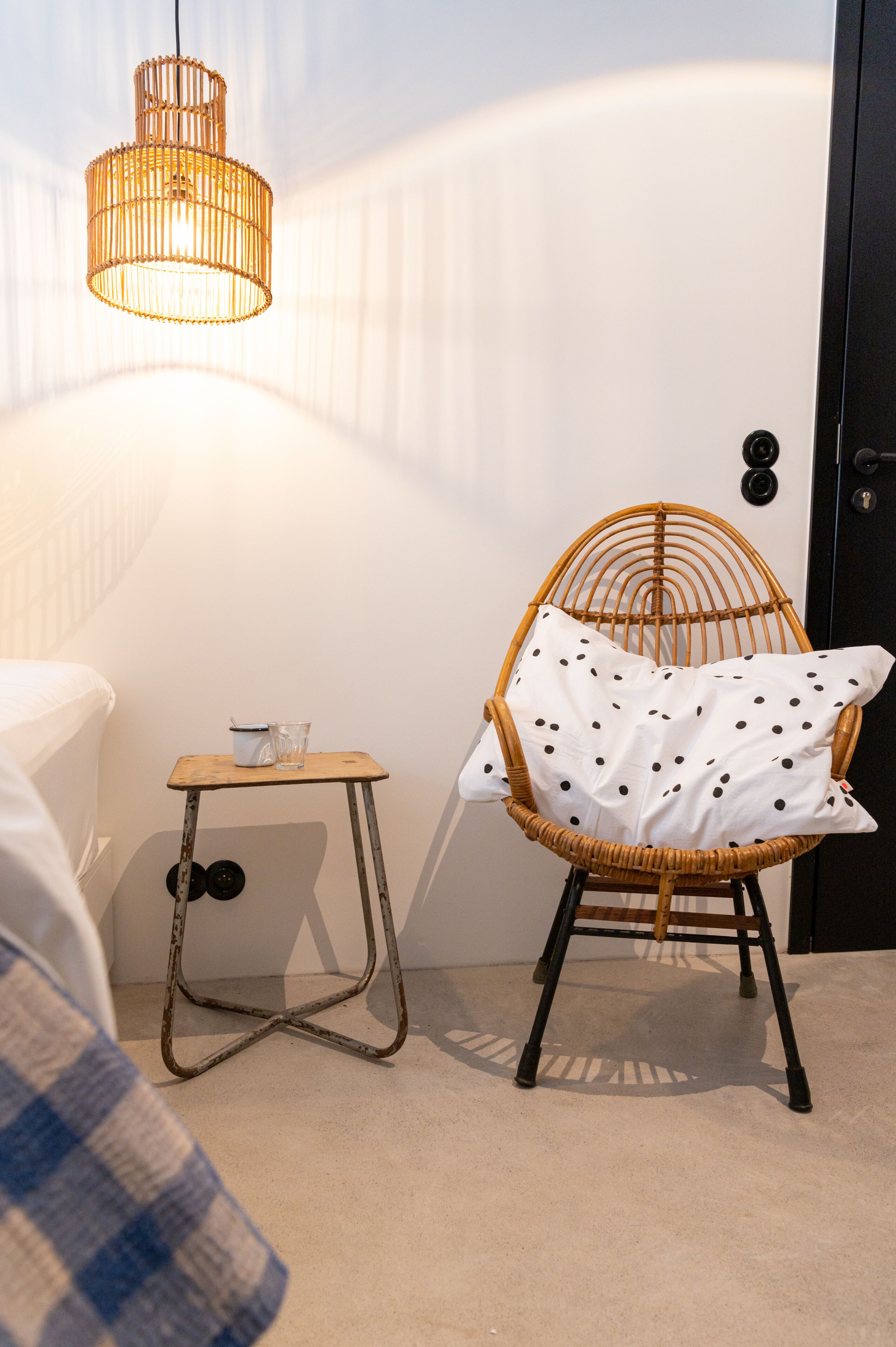 Ein Zimmer mit einem Korbstuhl, einem kleinen Holztisch mit einem Glas und einer Tasse sowie einer Hängelampe.