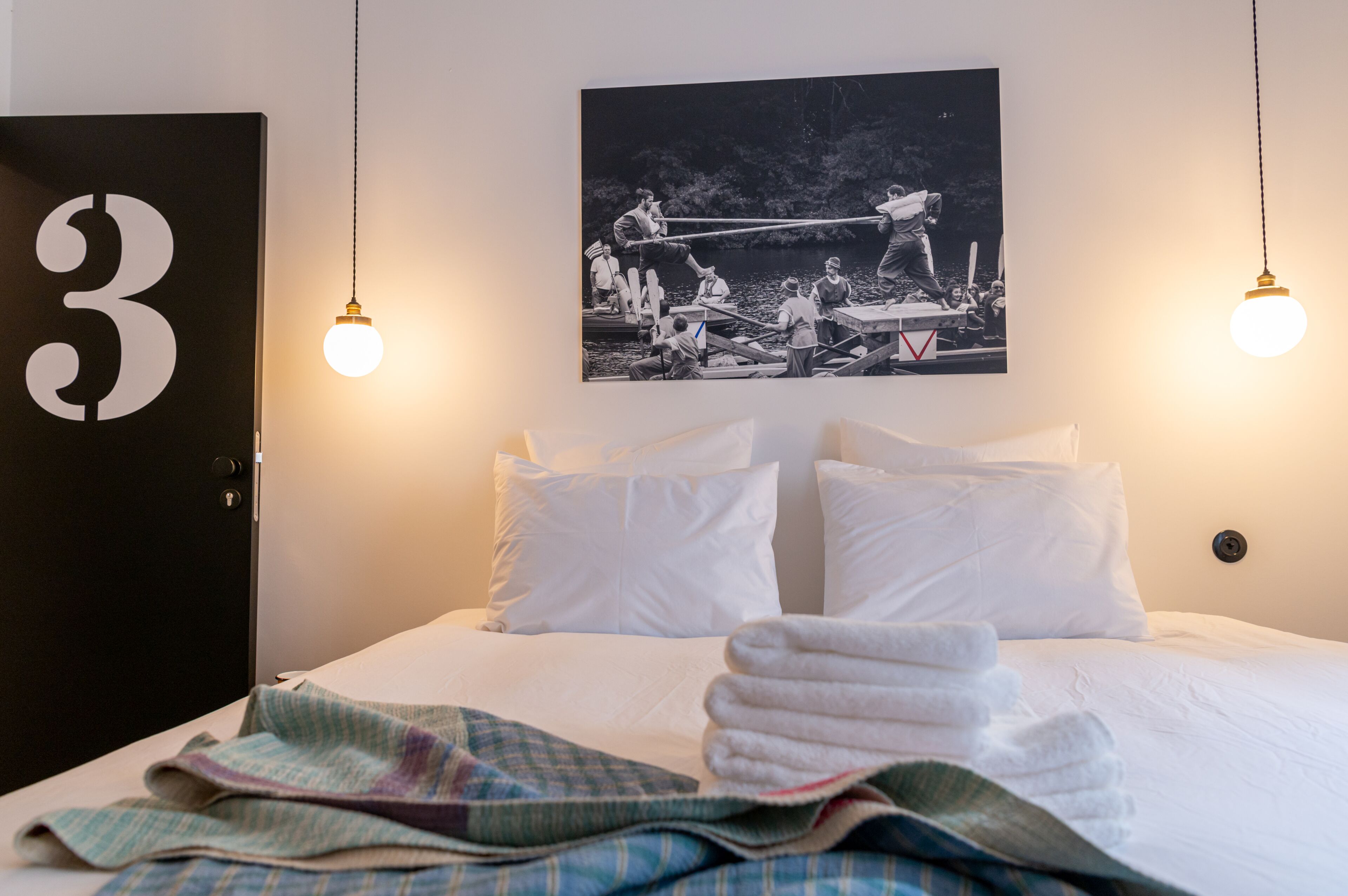 Ett hotellrum med en säng, vita kuddar, en svart dörr med nummer 3 och ett svartvitt fotografi på väggen.