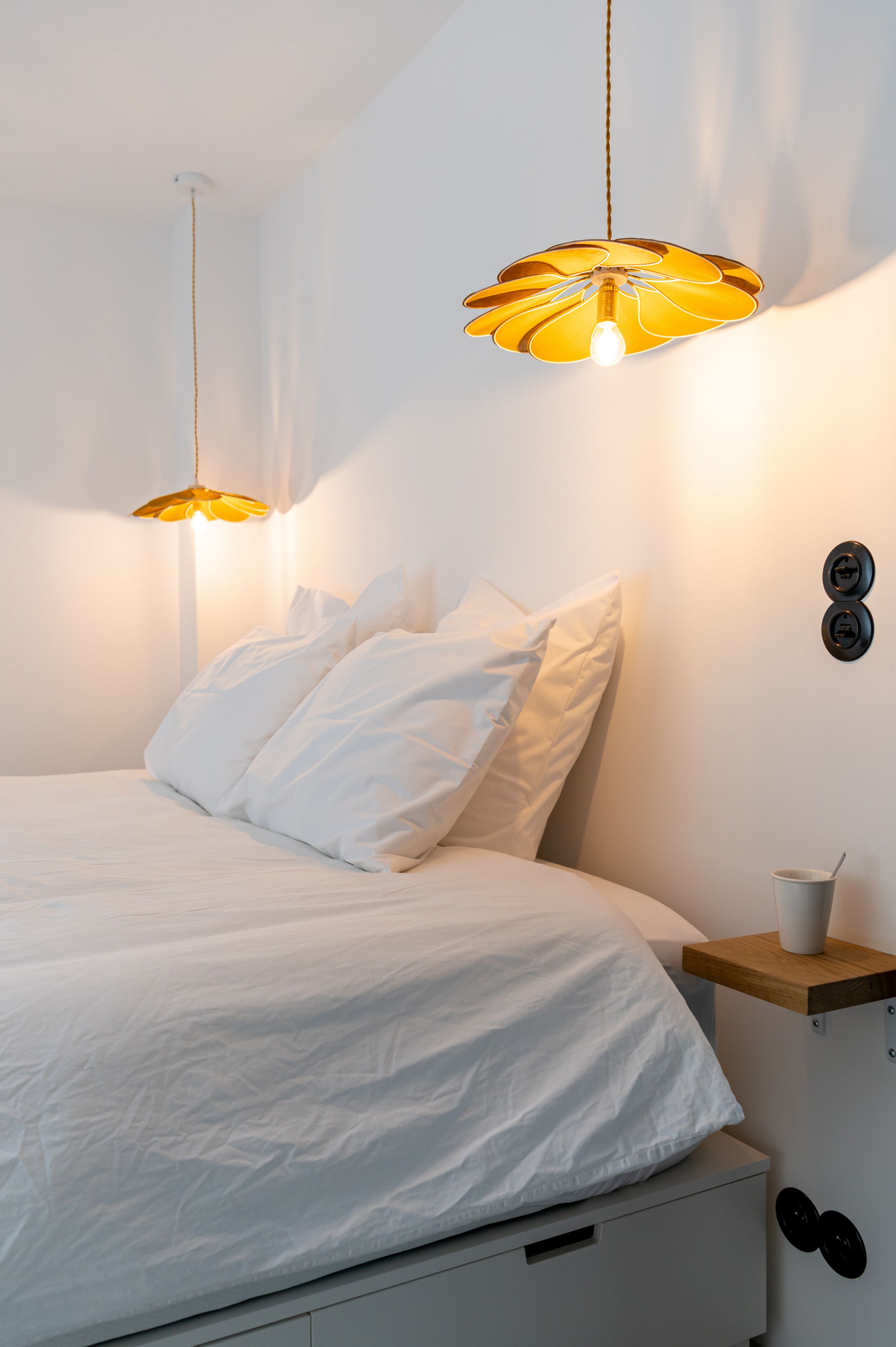 A bedroom with a bed, white bedding, two pendant lights, a bedside table with a cup, and a wall-mounted light switch.