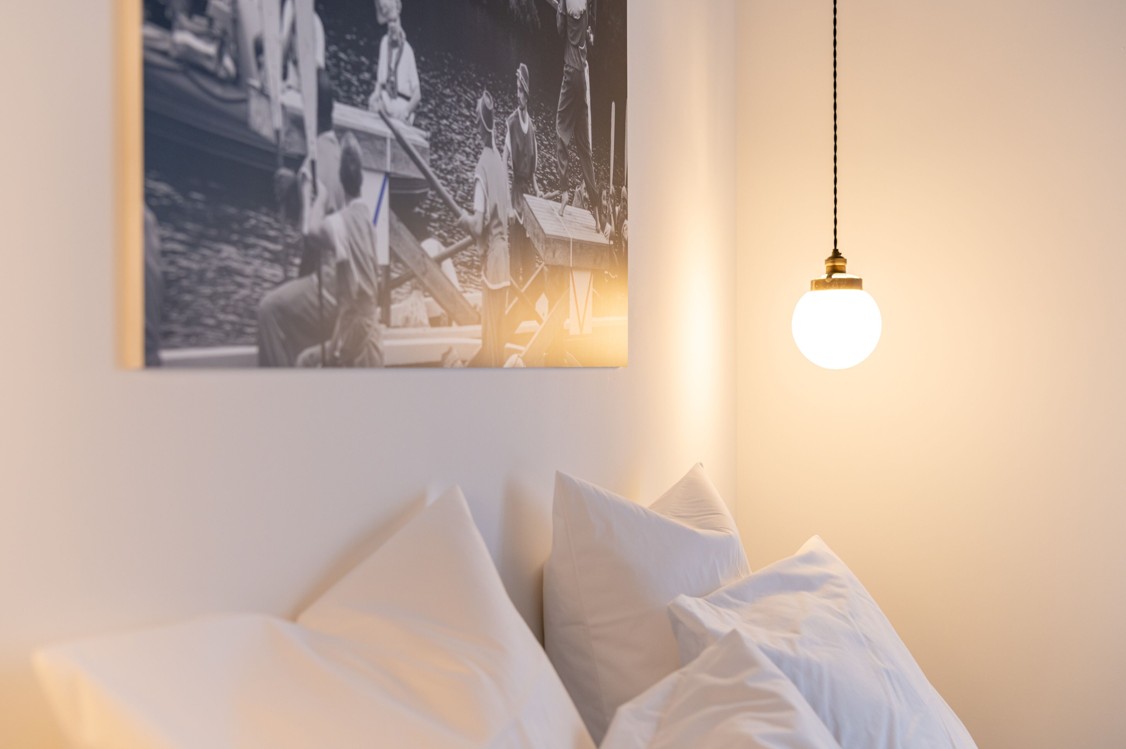 A bedroom with a bed, pillows, a hanging light fixture, and a black and white photograph on the wall.