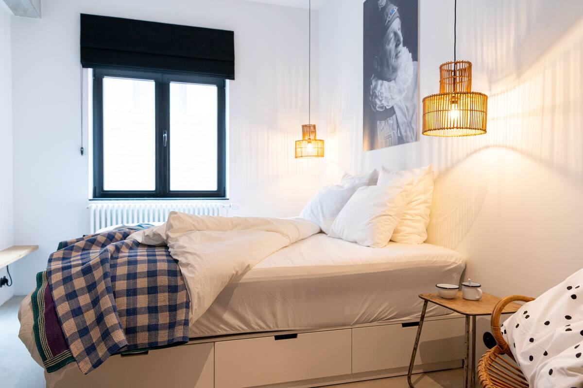 Een slaapkamer met een bed, een raam, een radiator, een wandlamp en een klein tafeltje met een stoel.