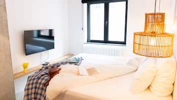 A modern hotel room with a bed, a flat-screen TV, a window with blinds, a bedside lamp, and a woven light fixture.